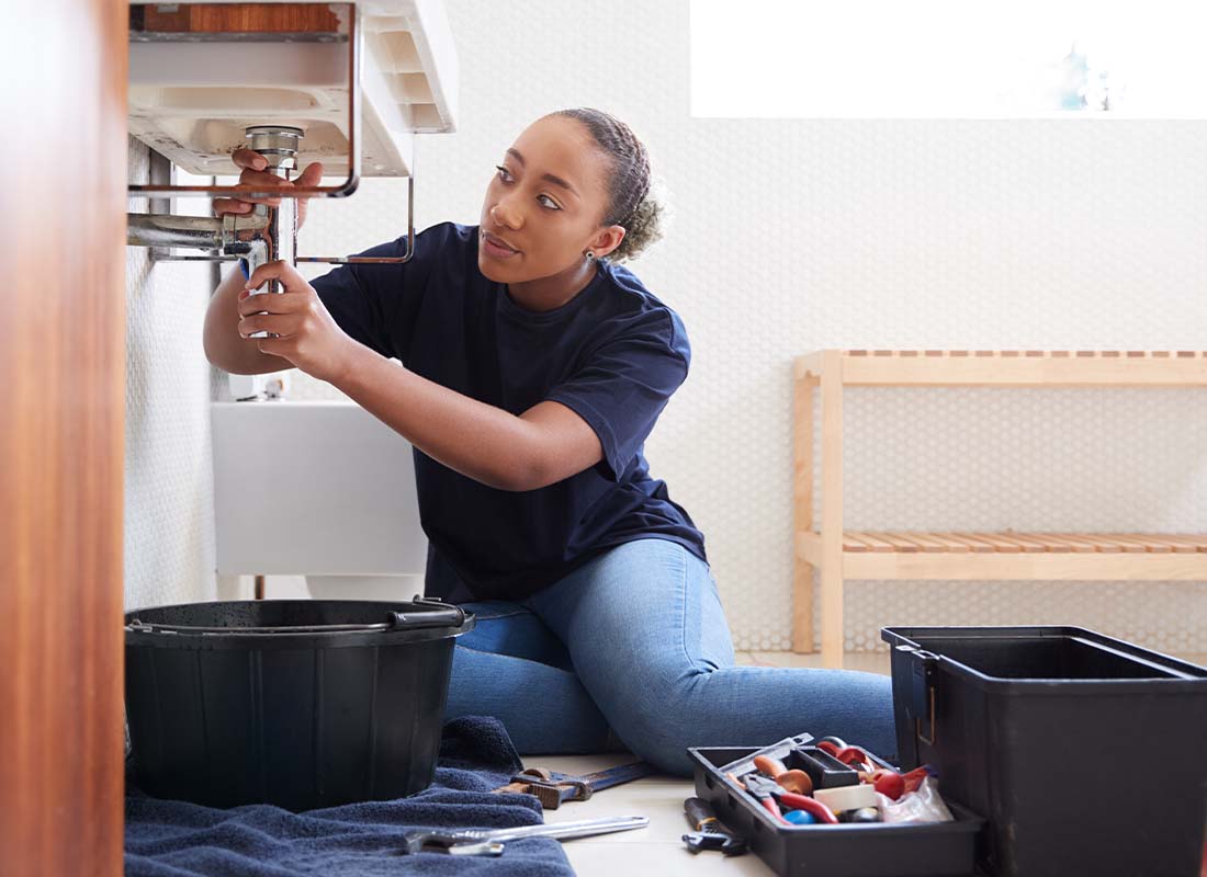 Plumbing Contractor Insurance - Female Plumber Sitting on the Floor and Working to Fix Leaking Sink In a Home Bathroom Seen From the Doorway